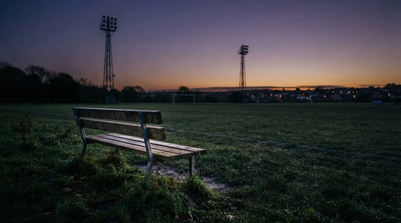 Eine leere Parkbank am Rand eines Fußballfelds in der Abenddämmerung, im Hintergrund ragen dunkle Flutlichtmasten in den Himmel.